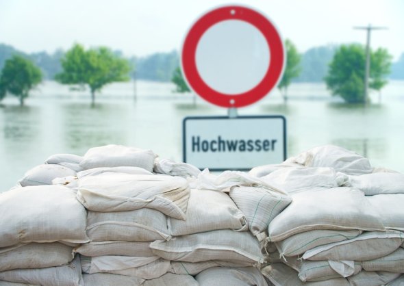 Symbolbild Hochwasser Schild mit der Aufschrift Hochwasser steht hinter gestapelten Sandsäcken. Im Hintergrund ist Wasser zu sehen.