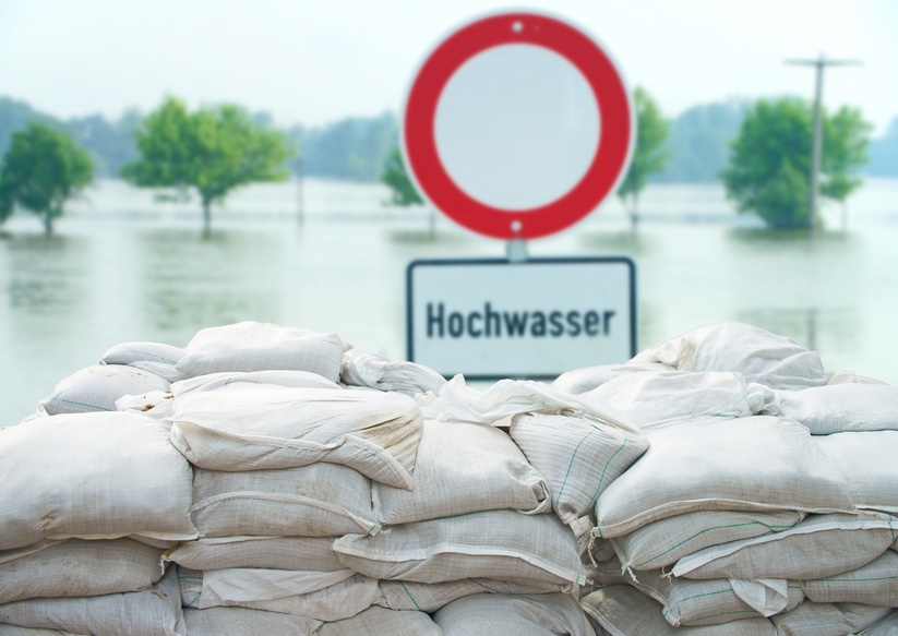 Schild mit der Aufschrift Hochwasser steht hinter gestapelten Sandsäcken. Im Hintergrund ist Wasser zu sehen. 