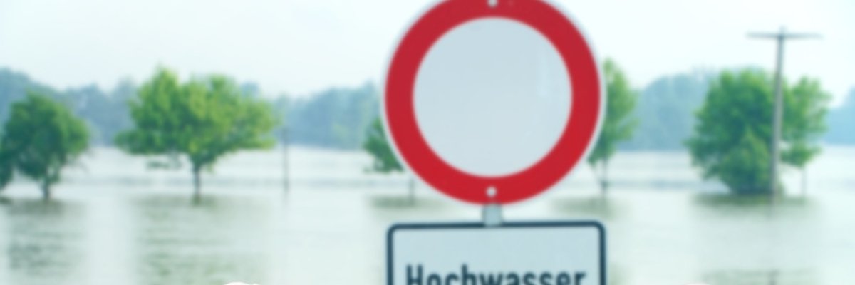 Schild mit der Aufschrift Hochwasser steht hinter gestapelten Sandsäcken. Im Hintergrund ist Wasser zu sehen. 