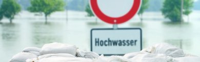 Schild mit der Aufschrift Hochwasser steht hinter gestapelten Sandsäcken. Im Hintergrund ist Wasser zu sehen. 