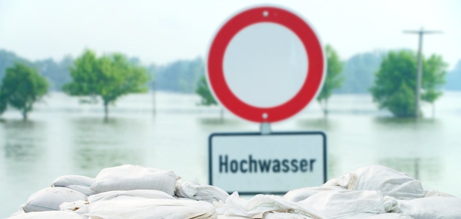 Symbolbild Hochwasser Schild mit der Aufschrift Hochwasser steht hinter gestapelten Sandsäcken. Im Hintergrund ist Wasser zu sehen.