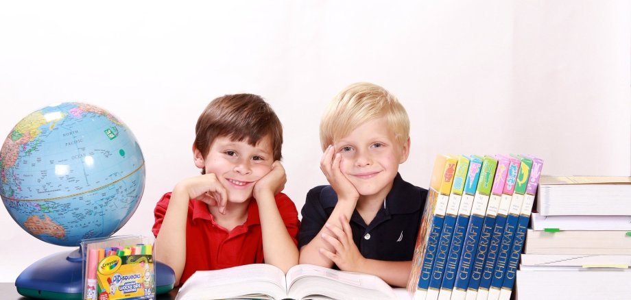 Jungen mit Büchern Zwei Jungs im Grundschulalter sitze an einem Tisch und lächeln in die Kamera. AUf dem Tisch steht ein Globus, Schulutensilien und ein Stapel mit Büchern.
