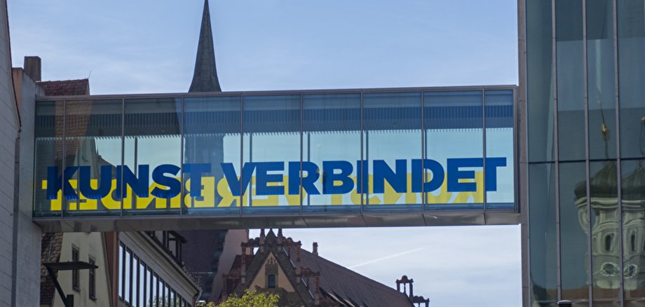 Kunst verbindet es wird eine Glasbrücke mit der Aufschrift "Kunst verbindet" gezeigt