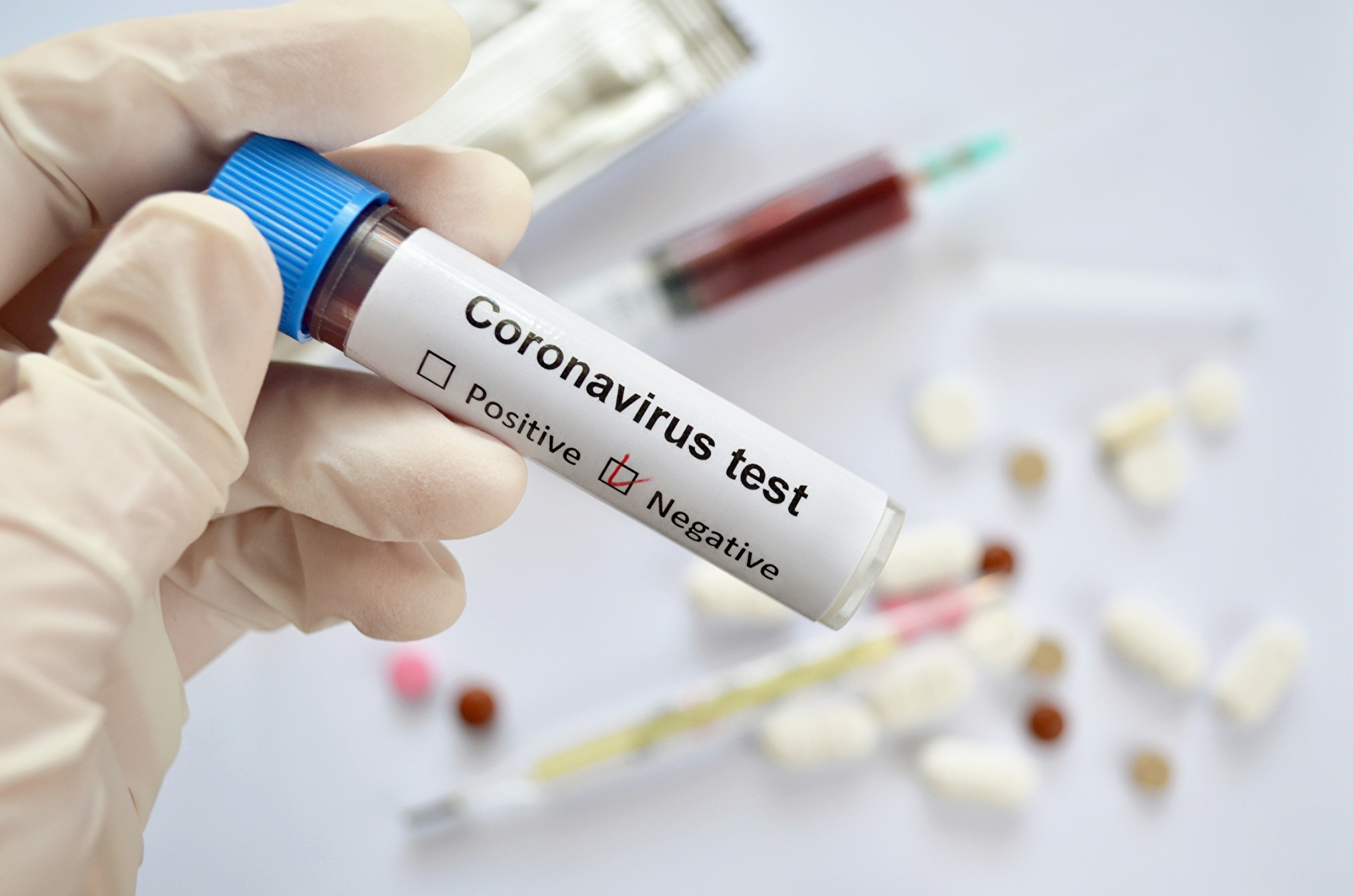 Reagenzglas mit Blutprobe und negativem Testergebnis für eine Coronavirus-Infektion in der Hand eines Arztes in Gummihandschuhen auf weißem, defokussiertem Hintergrund mit Medikamenten, Nahaufnahme