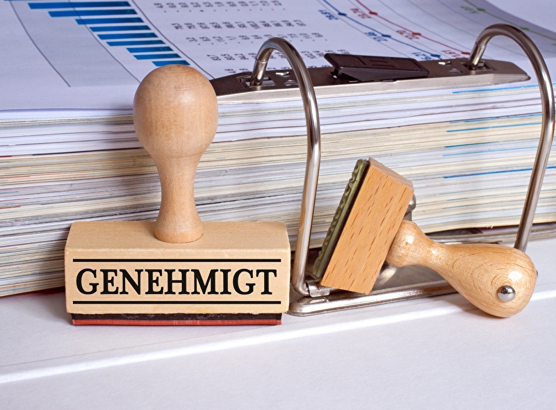 Stempel mit der Aufschrift Genehmigt steht vor einem aufgeschlagenen Aktenordner