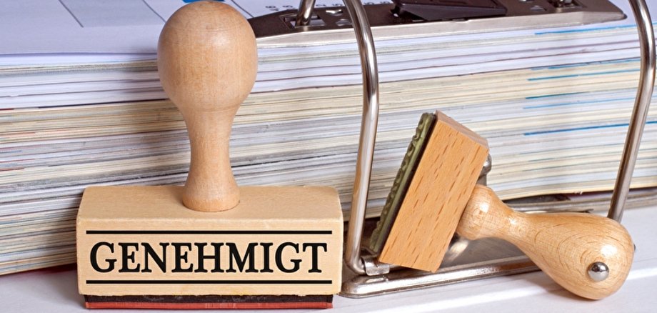 Symbolbild Genehmigung Stempel mit der Aufschrift Genehmigt steht vor einem aufgeschlagenen Aktenordner