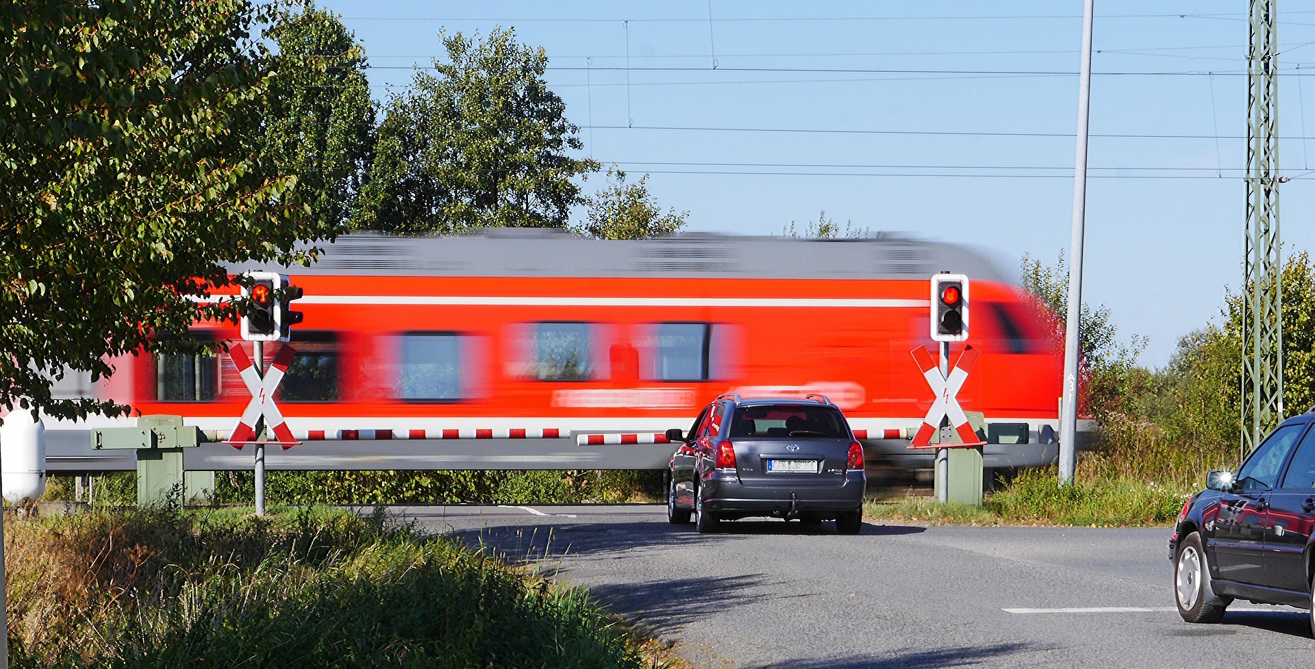 Ein roter Zug fährt über einen höhengleichen Bahnübergang. Ein Auto wartet an der geschlossenen Schranke.