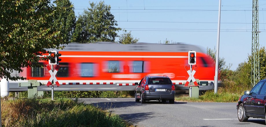 Ein roter Zug fährt über einen höhengleichen Bahnübergang. Ein Auto wartet an der geschlossenen Schranke.
