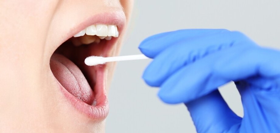 Symbolbild Corona-Test Nahaufnahme. Eine Hand in einem blauen Latexhandschuh nimmt einen Rachenabstrich bei einer Person mit geöffnetem Mund.