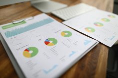 Symbolbild Finanzbericht Nahaufnahme eines Finanzberichtes auf einem Holztisch. Auf den zwei Blättern sind verschiedene Diagramme und Statistiken zu sehen. Im Hintergrund sieht man eine Tastatur.